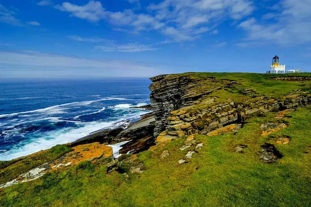 A white lighthouse on green clifftops in Orkney above a blue Atlantic swell